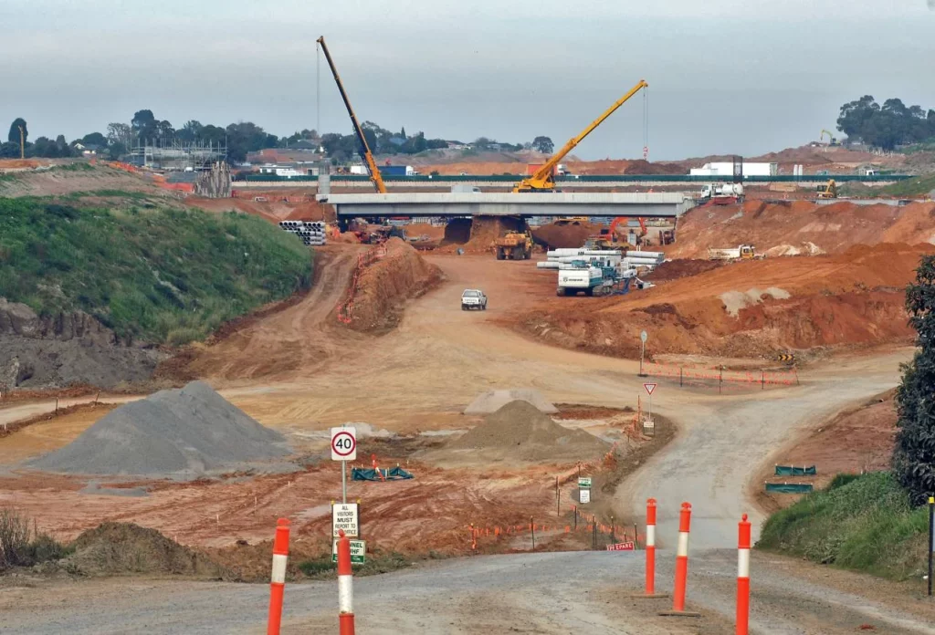 A large construction site with cranes, trucks, and equipment building a bridge over a wide, cleared area of dirt and gravel. Orange cones, piles of earth, and construction signs line the road in the foreground.