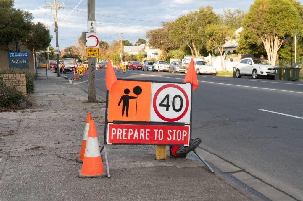 A roadside sign displays a 40 km/h speed limit and "PREPARE TO STOP," with orange cones and traffic in the background, indicating nearby roadwork on a suburban street.
