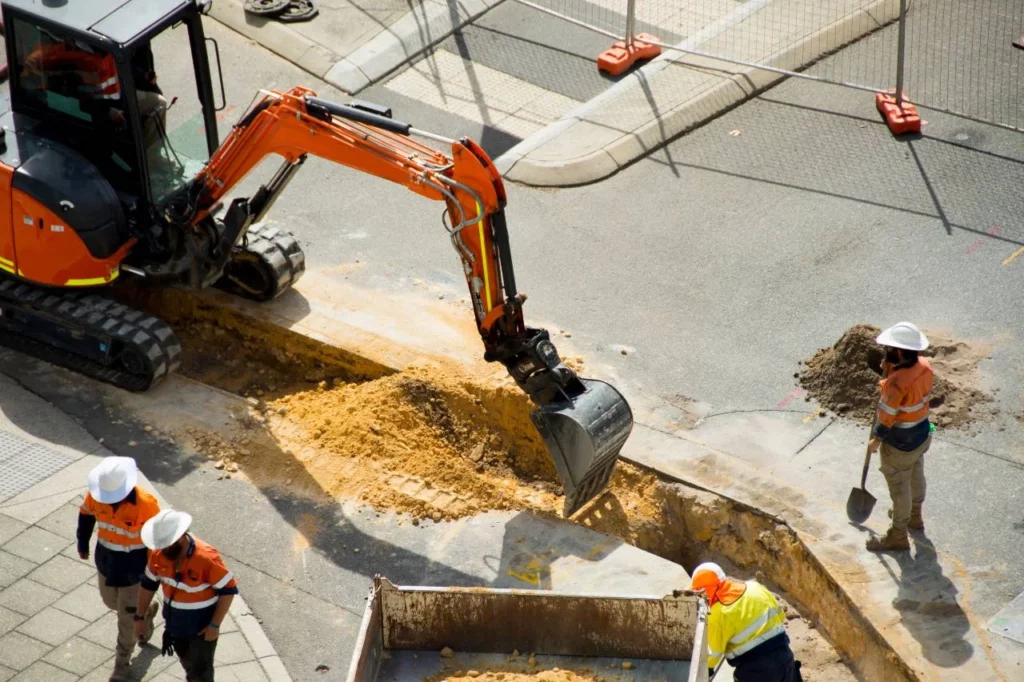 A construction crew works on a city street, using an orange excavator to dig a trench. Three workers in safety gear stand nearby, with one shoveling dirt and another near construction barriers.
