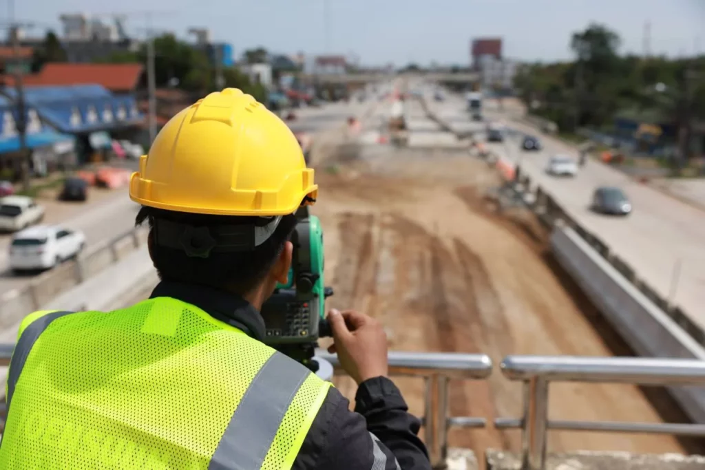 A construction worker wearing a yellow hard hat and neon safety vest uses surveying equipment to measure and observe a large road construction site with traffic and buildings in the background.