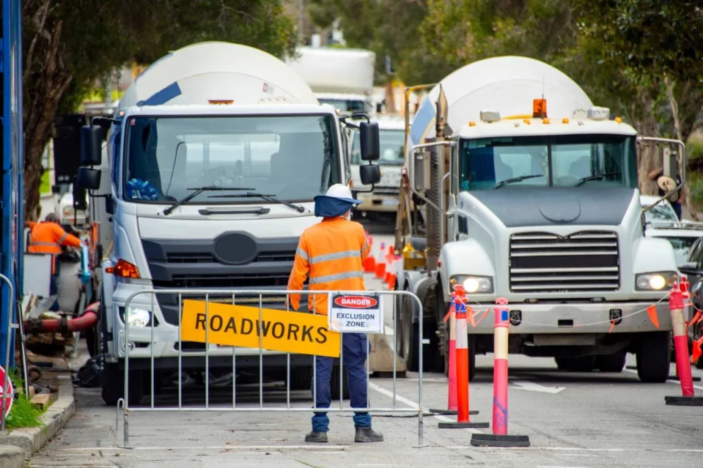 A construction worker in a high-visibility jacket stands in front of a "ROADWORKS" sign and safety barrier, blocking two large trucks on a street lined with traffic cones and trees.