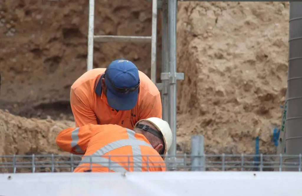 Two construction workers wearing orange safety jackets and helmets are working together at a construction site with a dirt wall in the background.