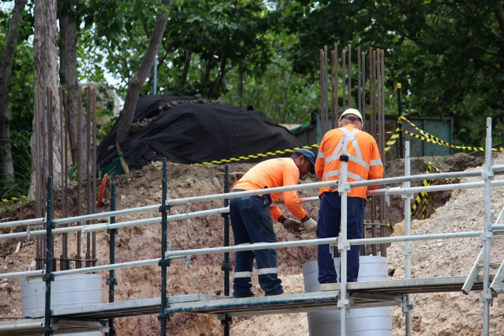 Two construction workers in orange high-visibility shirts work on a scaffolding structure at a building site. Rebar and safety tape are visible, with trees and soil in the background.
