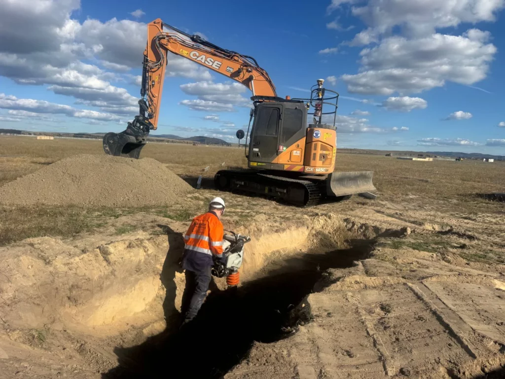 A construction worker in a high-visibility outfit operates a compactor in a trench, while an orange CASE excavator is parked nearby on a wide, open, grassy field under a blue sky with clouds.