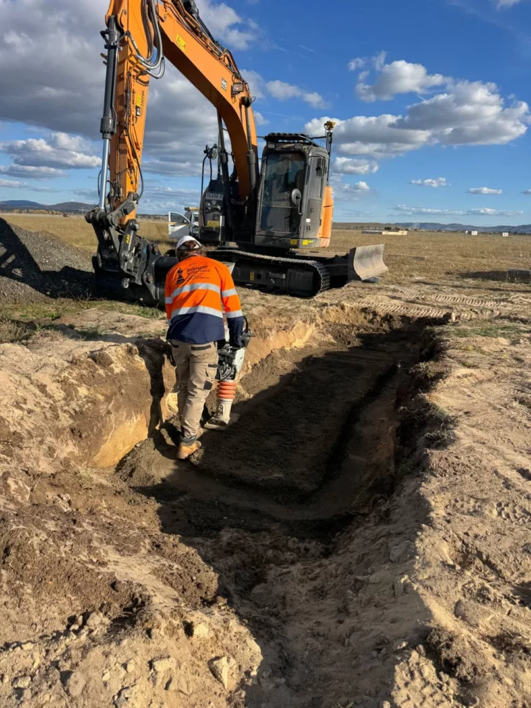 A construction worker in a high-visibility orange jacket and helmet stands in a freshly dug trench on a dirt site, with an excavator parked nearby under a partly cloudy sky.