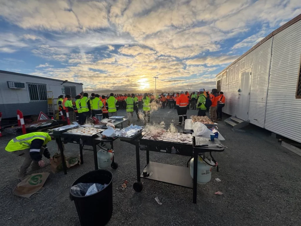 A group of construction workers in high-visibility vests gather near trailers at sunrise or sunset, with food and grills set up on tables in the foreground, suggesting a meal or event at a worksite.