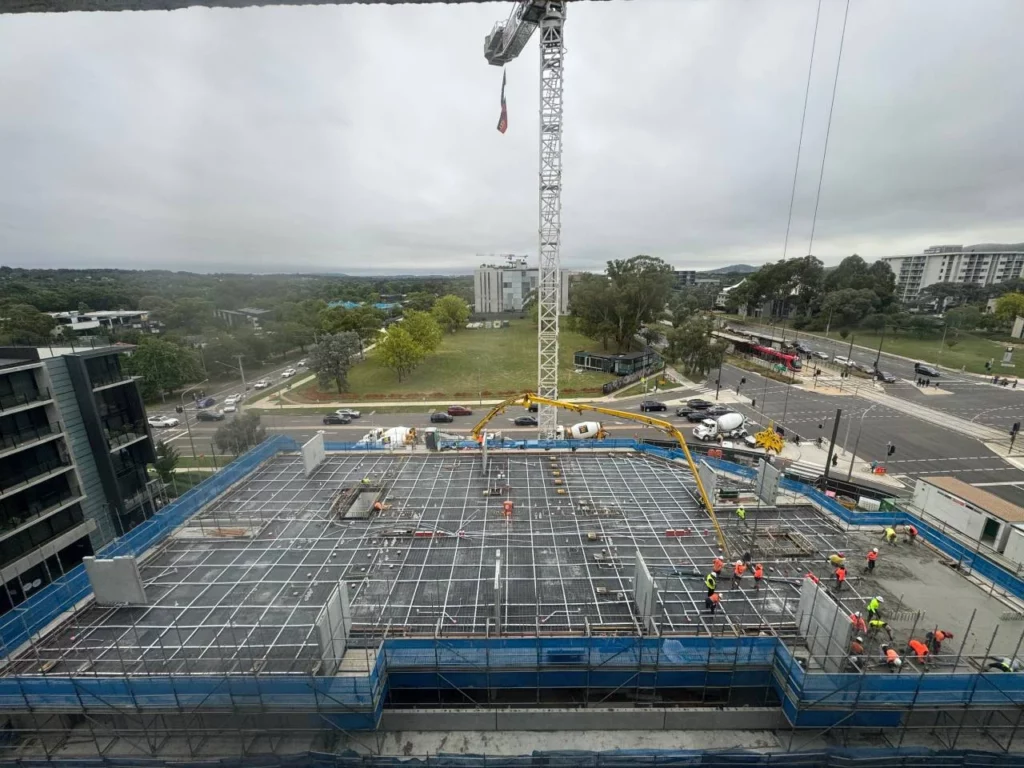 A group of construction workers on a building site with a concrete slab framework, surrounded by blue safety barriers. A crane and concrete pump are visible, with roads, trees, and buildings in the background under a cloudy sky.