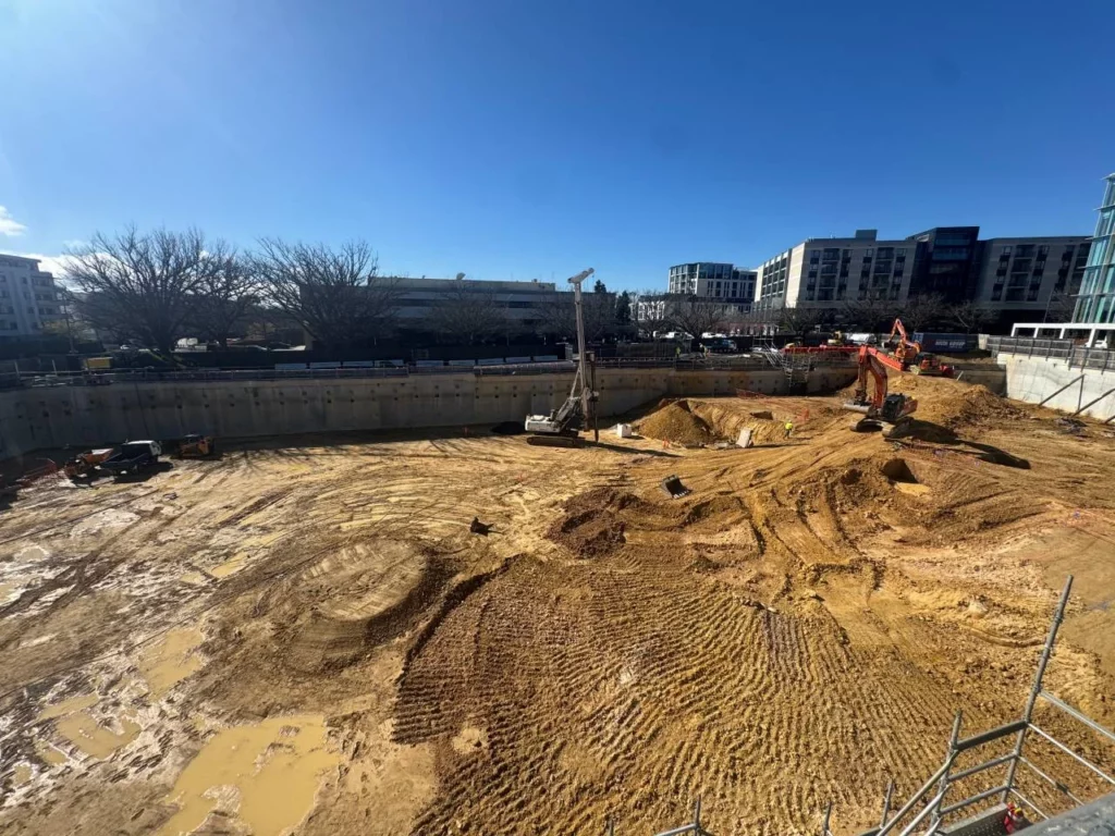 A construction site with excavation machinery on muddy ground, surrounded by deep retaining walls. Buildings and leafless trees are visible in the background under a clear blue sky.