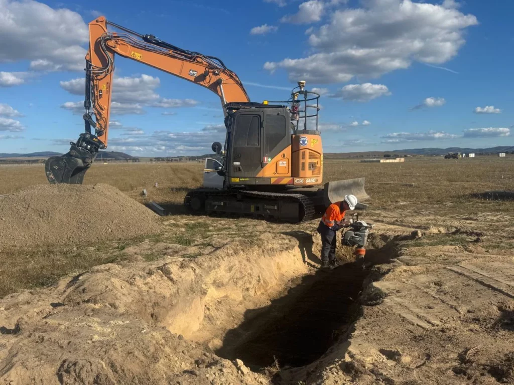 A construction worker in safety gear operates machinery beside a large trench, with an excavator and a pile of dirt nearby, in an open field under a blue sky with scattered clouds.